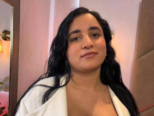 LisaStarkm with long black wavy hair, wearing a white robe, has a medium skin tone, brown eyes, and a slight smile, showing a small pimple on her right cheek.