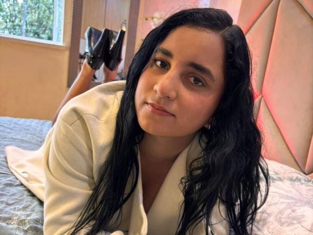 LisaStarkm, allongée sur le lit, portant une robe blanche, tenant des escarpins noirs, cheveux noirs ondulés, peau claire, maquillage discret, léger sourire, la tête reposant sur la main, lit avec des draps à motifs..