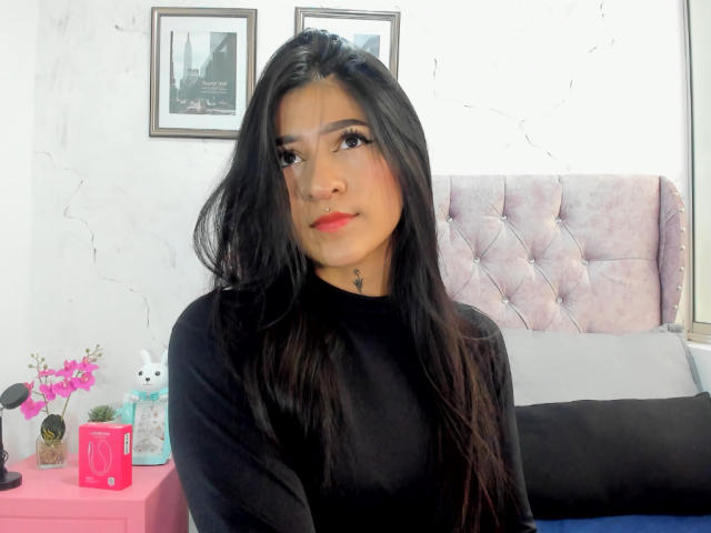 LuciadelaVega with long black hair, black top, red lipstick, and nose ring, sitting in front of a pink tufted headboard, looking at the camera, with a small pink table.