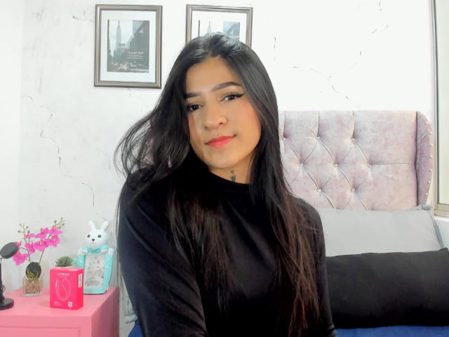 LuciadelaVega with long black hair, wearing a black shirt, sitting on a bed, smiling, cute, pink nose, bedroom with pink bed, white wall, and framed pictures.