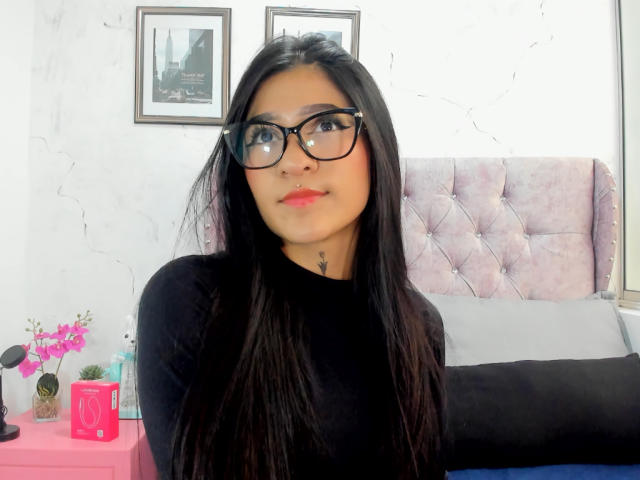 LuciadelaVega with long black hair, black glasses, red lipstick, black top, and nose ring, sitting in front of a pink tufted headboard, looking up with a serious expression.