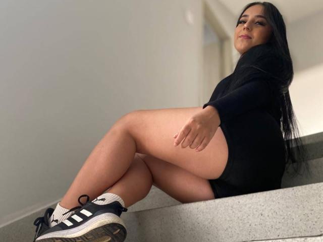 StefanyDelly with long black hair, wearing a black long-sleeve top and black shorts, sitting on a grey staircase, legs crossed, showing her ass, in black and white Adidas sneakers.