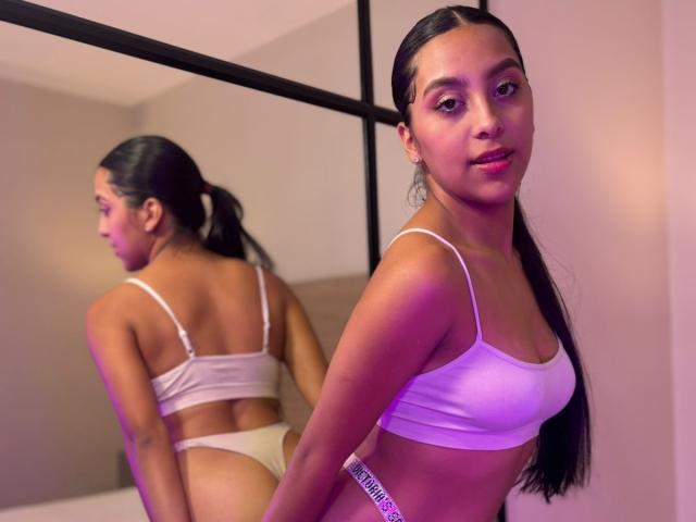 EmmaEstreilla with long black hair in a ponytail, wearing a white bra and panties, showing her back in a mirror. Her small tits and cute ass are visible.
