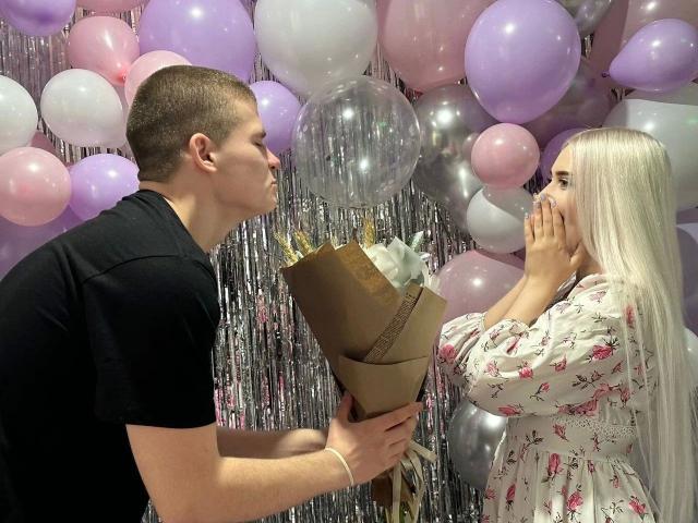 KevinKRYSTALLL-hot, short brown hair, black shirt, leans in with brown bouquet to long-haired KevinKRYSTALLL-hot, pink floral dress, hands over mouth, pastel balloons, silver streamers.