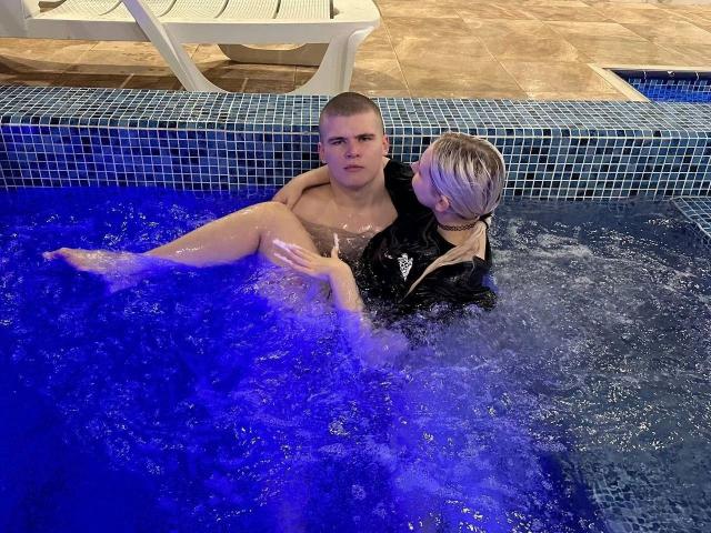 KevinKRYSTALLL-hot with a shaved head, shirtless, sitting in a blue-tiled pool with blue water, holding another KevinKRYSTALLL-hot with blonde hair, black jacket, and visible ass.