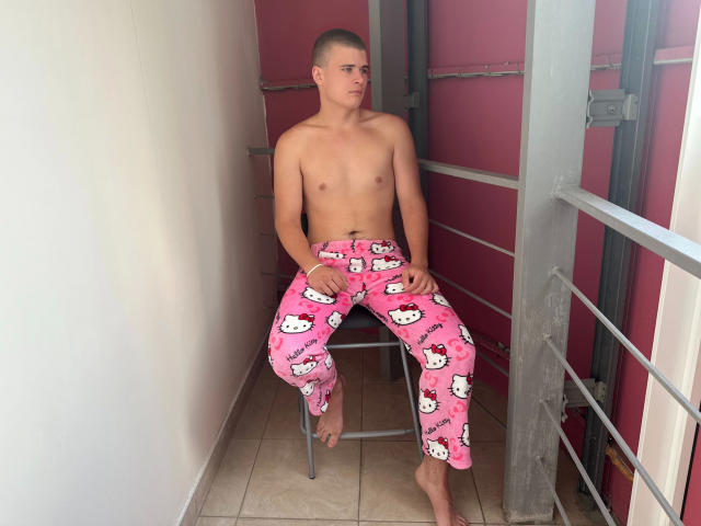 KevinKRYSTALLL-hot, shirtless, with shaved head, wearing pink Hello Kitty pajama pants, sitting on a chair, looking to the side, bare feet on tiled floor, in a narrow hallway.