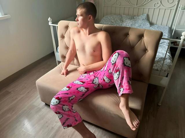 KevinKRYSTALLL-hot, shirtless with pink Hello Kitty pajamas, sitting on a beige tufted couch, legs spread, bare feet, short hair, bed in background, wooden floor.