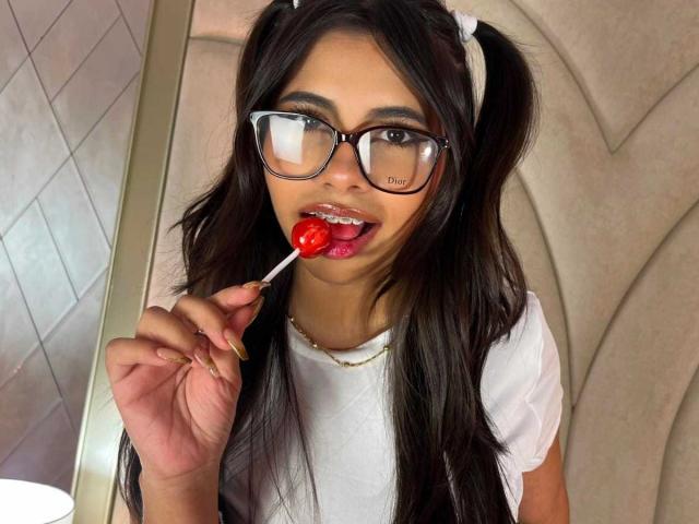 Celeste Morel, con lunghi capelli neri raccolti in due codini, indossa occhiali e una camicia bianca, e sta leccando un lecca-lecca rosso con la lingua fuori. Le sue natiche non sono visibili..
