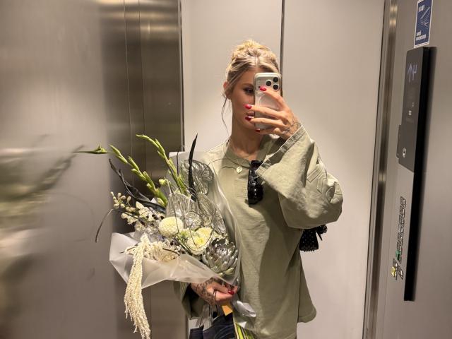 SensualMila with blonde hair in a bun, wearing a green shirt, holding a bouquet of flowers, taking a mirror selfie in an elevator, showing off her red nails.
