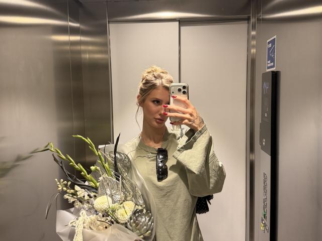 SensualMila with blonde hair in a bun, wearing a green shirt, holding a bouquet of white and green flowers, taking a mirror selfie in a metallic elevator.