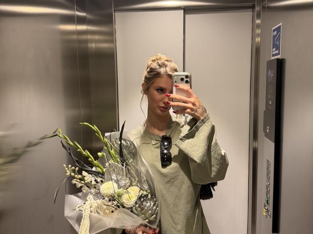 SensualMila with blonde hair in a bun, wearing a green shirt, holding flowers, taking a mirror selfie in an elevator, red nails, black sunglasses in hand.