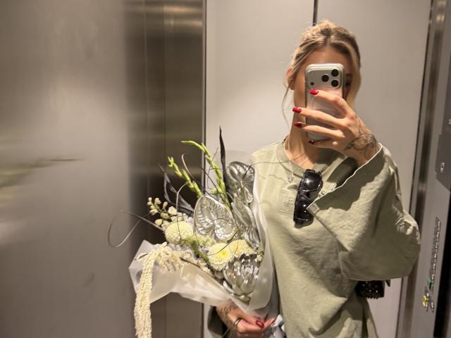 SensualMila, avec des cheveux blonds, portant une robe verte, tenant un bouquet de fleurs, selfie pris dans un miroir, ongles rouges, tatouage sur la main droite, smartphone, lunettes de soleil sur la poitrine, ascenseur..