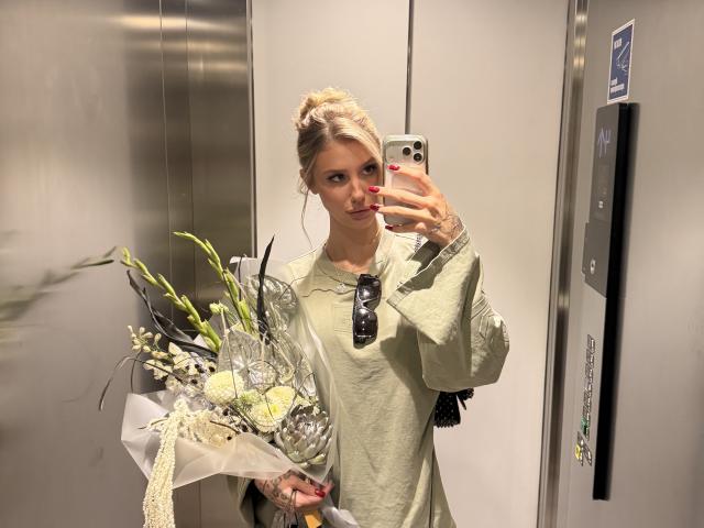 SensualMila with blonde hair in a bun, holding a bouquet of flowers, wearing a light green shirt, taking a mirror selfie in an elevator, red nails, tattoos.