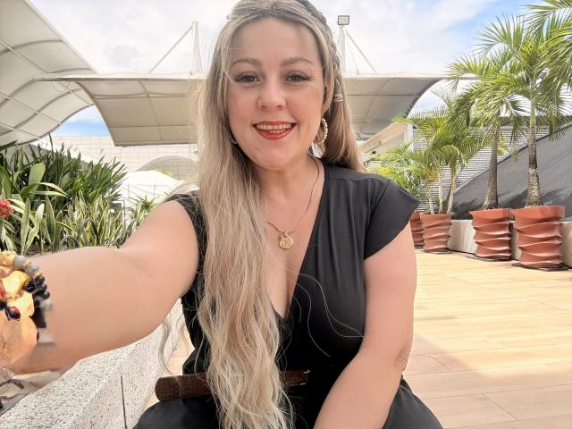 SamanthaGold, met lang blond haar, lachend, gekleed in een zwarte jurk, met een gouden ketting en armband, steekt haar arm uit voor een selfie, waarbij haar borsten licht zichtbaar zijn, buiten, zonnig..