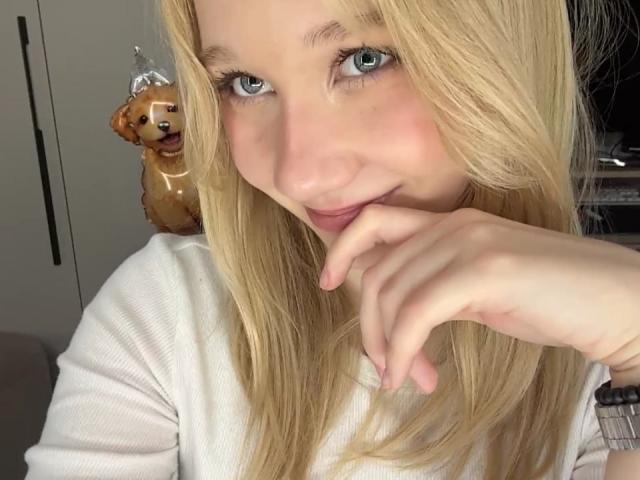 Ruth Demerson hat blondes Haar, blaue Augen und lächelt, wobei sie mit ihrem Finger in der Nähe ihres Mundes ist. Sie trägt ein weißes Hemd, und hinter ihr befindet sich ein süßes Hundespielzeug mit einer Krone..