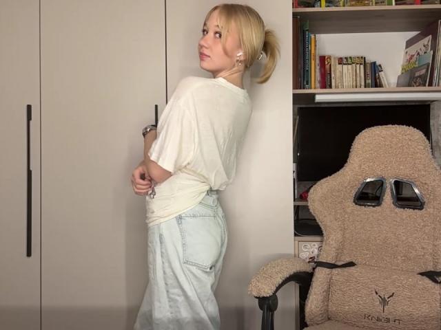 RuthDemerson with blonde ponytail, white tee, light blue jeans, looking over shoulder, standing, beige room, beige chair with "RAGE" logo, bookshelf, and white cabinet in background.