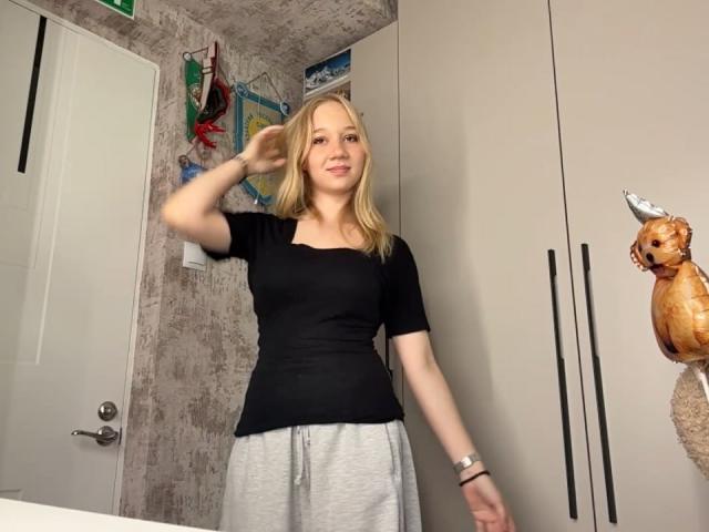 Ruth Demerson, blond, schwarzes Oberteil, graue Hose, steht in einem Raum, rechter Arm erhoben, lächelnd, weiße Tür, Wand mit Karte, beigefarbene Schränke, kleines Stofftier auf der rechten Seite..
