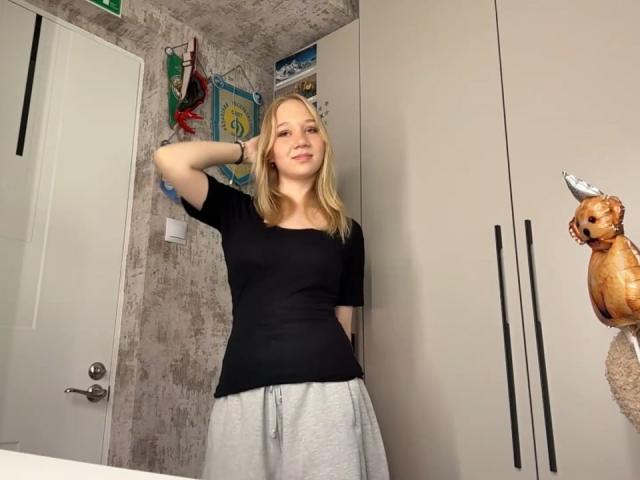 Ruth Demerson steht in einem Raum, trägt ein schwarzes T-Shirt und graue Hosen, hat blondes Haar, einen Arm angehoben und lächelt entspannt, neben einem Stoff-Eichhörnchen..