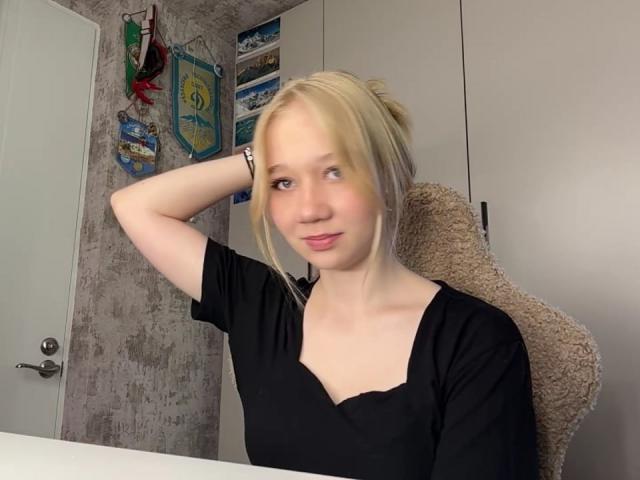 Ruth Demerson mit blonden Haaren zu einem Dutt gebunden, trägt ein schwarzes Oberteil, lächelt leicht, hat einen Arm angehoben und sitzt auf einem beigen Stuhl. Im Hintergrund sind eine weiße Tür und eine weiße Wand zu sehen..