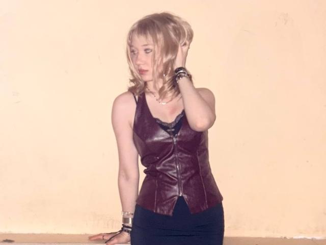 Ruth Demerson, avec ses cheveux blonds, portant un gilet en cuir violet, une jupe noire et des bracelets, touchant ses cheveux, montrant sa petite poitrine et ses hanches fines. Elle regarde vers le bas..