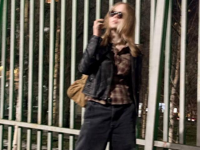 Ruth Demerson mit langem, blondem Haar, Sonnenbrille, schwarzer Lederjacke, kariertem braunem Hemd, schwarzen Hosen, einer beigen Tasche, telefoniert und steht nachts neben grünen Metallstreben..