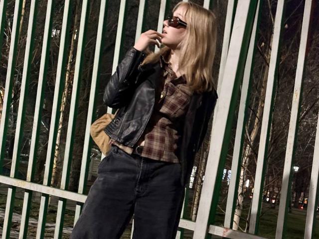 Ruth Demerson mit blonden Haaren, einer schwarzen Lederjacke, einem braun-karierten Hemd, schwarzen Hosen und einer Sonnenbrille, steht vor grünen Metallstreben, hält ein Telefon ans Ohr und trägt eine braune Tasche über der Schulter..