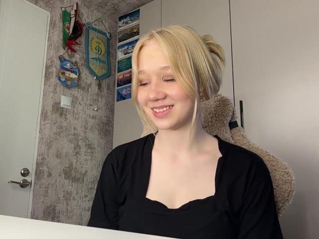 RuthDemerson, blonde with fair skin, smiling with eyes closed, wearing a black shirt, sitting in a chair, her small tits slightly visible, in a brightly lit room.