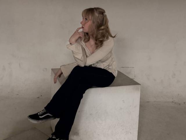 Ruth Demerson, met blond haar, zittend op een witte kubus, gekleed in een beige top, zwarte broek en zwarte sneakers, denkt met haar vinger in de buurt van haar mond..