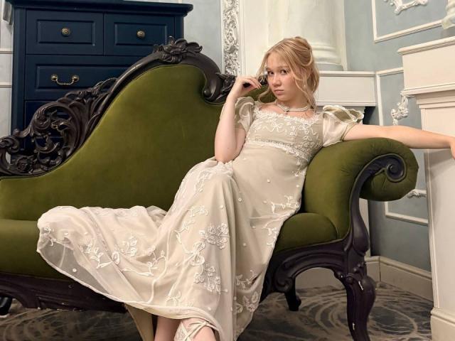 Ruth Demerson, blonde, vêtue d'une robe blanche en dentelle ornée de motifs floraux, est assise sur un vieux canapé vert en velours, parlant au téléphone, et dégage une aura détendue, séduisante et élégante..