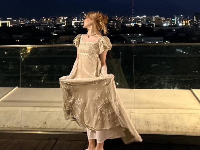 RuthDemerson está em um telhado à noite, vestindo um vestido bege claro com bordados florais, segurando a saia. Ela tem uma estrutura esguia, cabelo loiro e um colar..