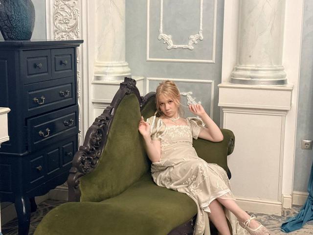 Ruth Demerson mit blonden Haaren, in einem transparenten weißen Kleid, sitzt auf einer olivgrünen, verzierten Liege und hält ein blaues Glasornament, während sie in die Kamera schaut..