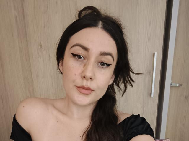 MabyLegrand has pale skin, dark brown hair in a messy ponytail, black off-shoulder top, winged eyeliner, and a small mole on her nose, looking at the camera.