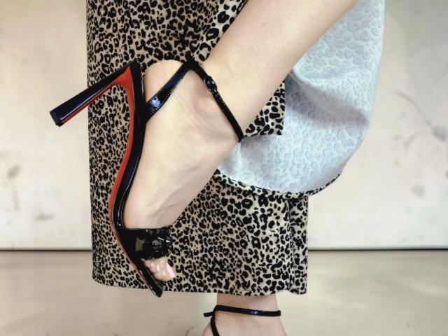 De voet van MabyLegrand in een zwarte stiletto met rode zolen, met een rok met luipaardprint en witte kant. Ze tilt haar been op, waardoor zwarte bandjes op haar enkel zichtbaar zijn..