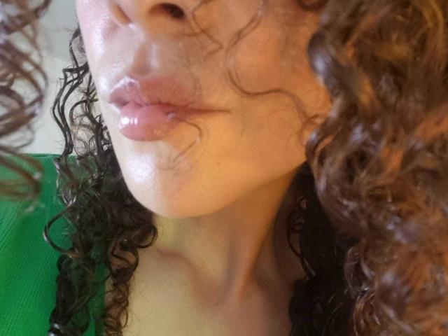 Le labbra piene di MissSunnyShine sono lucide, i suoi capelli castani ricci sono bagnati e indossa una maglietta verde, che mette in mostra il suo viso leggermente lucido e abbronzato..