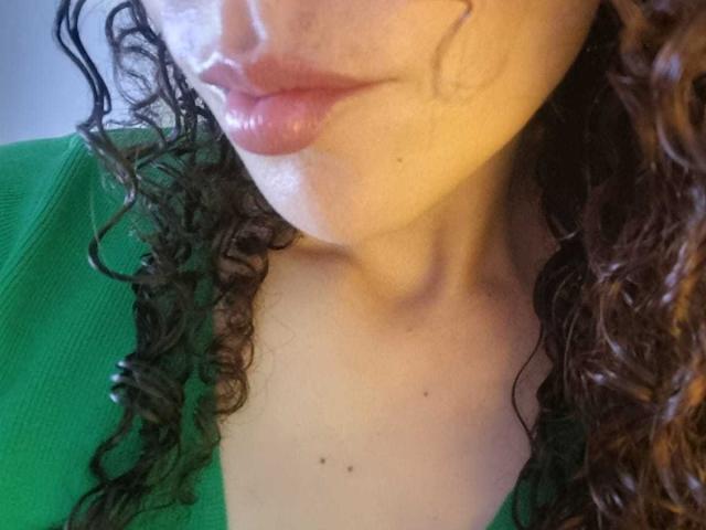 Il viso di MissSunnyShine, con le sue labbra rosa lucide, i capelli castani ricci e una maglietta verde, mostra la clavicola e una piccola lentiggine. La sua pelle è chiara e liscia..