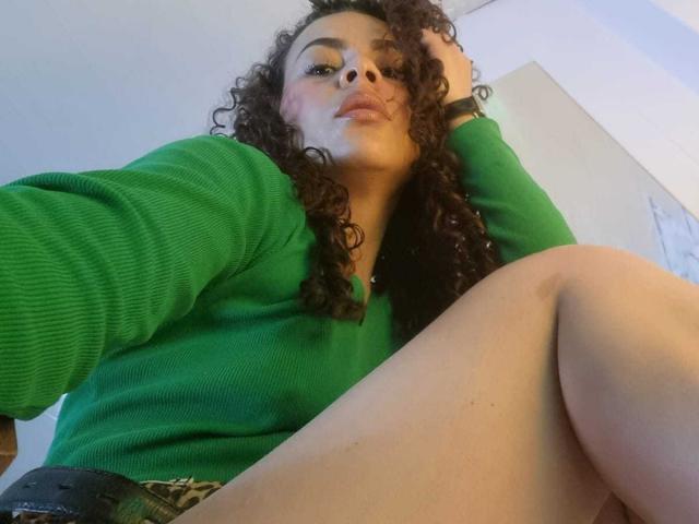 MissSunnyShine, con i capelli castano ricci, un maglione verde e in posa con le ginocchia leggermente sollevate, mostra il sedere e ha gli occhi verdi, guardando verso il basso la fotocamera. Le sue labbra sono leggermente tumefatte..