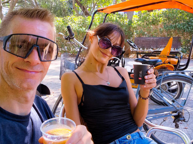 CassandraFredStyler and a dude with glasses, both smiling, sitting outside. CassandraFredStyler in black top, blue jeans, holding coffee, wearing sunglasses. Dude in black shirt, holding drink. Bikes.