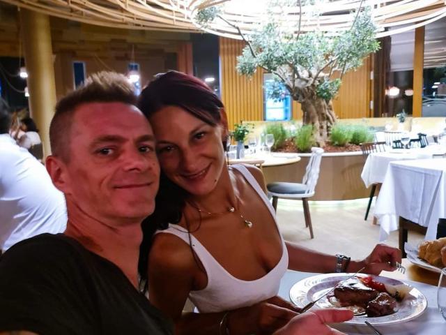 CassandraFredStyler with her tits pressed against a man's shoulder, smiling at the camera, both holding plates of food in a restaurant. She's wearing a white tank top.