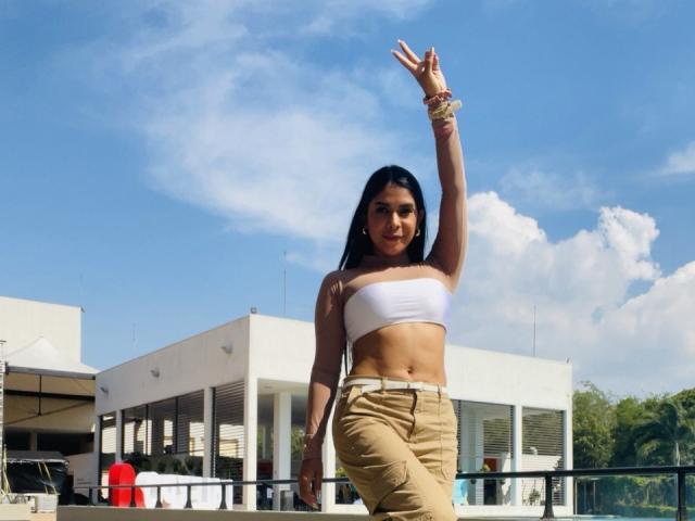 Mademoiselle, vestindo calças bege e um top branco, exibe seus músculos abdominais tonificados e levanta um braço com um sinal de paz, tem cabelos pretos longos e um sorriso confiante..