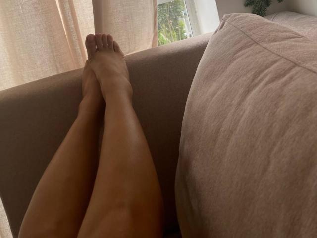 MademoiselleGo's bare legs crossed on a beige couch, feet with painted toenails, soft light, and a green plant partially visible in the background.