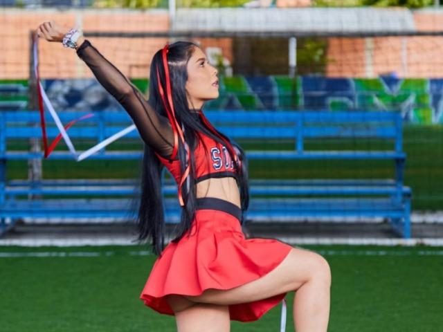 MademoiselleCom cabelos longos e pretos, uniforme de cheerleader vermelho, mangas de malha preta, braço levantado, segurando fitas, mostrando o bumbum, em pé em uma perna, ao ar livre, durante o dia, atlética, confiante..
