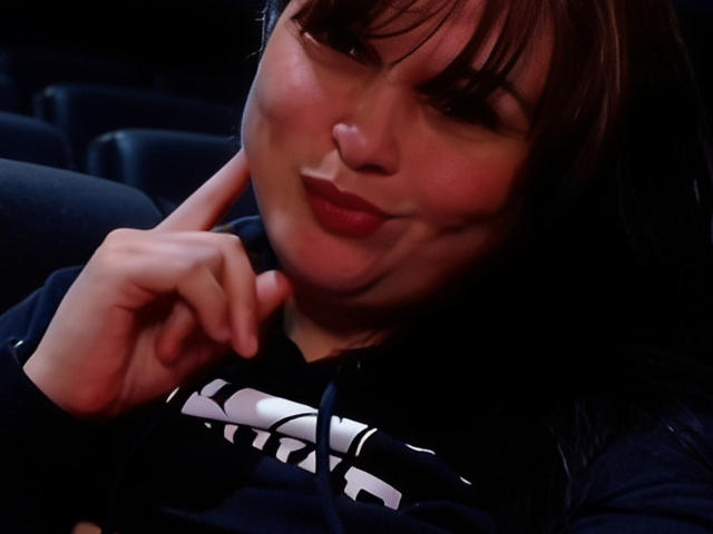 ValentineCurvy with pouty lips, brown hair, and bangs, wearing a black hoodie, resting her chin on her hand, smiling slightly, in a dimly lit, dark environment.