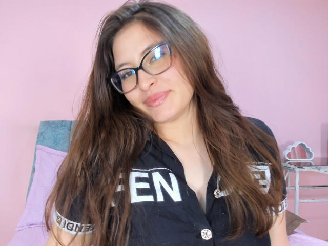 CassiBell with long brown hair, glasses, and a black shirt, showing off her tits, slight smile, pink background, and a silver necklace.