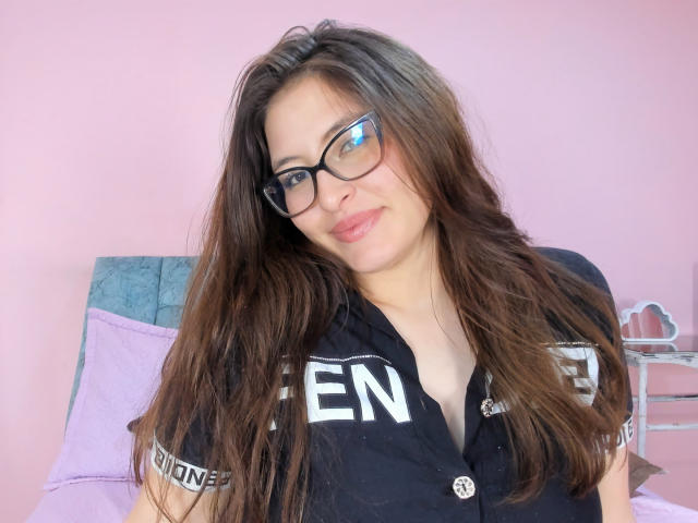 CassiBell with long brown hair, glasses, and a black shirt, showing a subtle smile, her tits slightly visible under the shirt. Pink wall and bed in the background.
