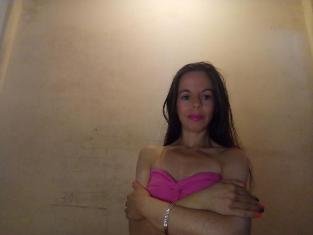 LaraF con el cabello castaño mojado, un sujetador rosa sin tirantes, los brazos cruzados, esmalte de uñas rojo, una pulsera de plata y lápiz labial rosa, de pie en una habitación con poca luz. Se ven los senos..