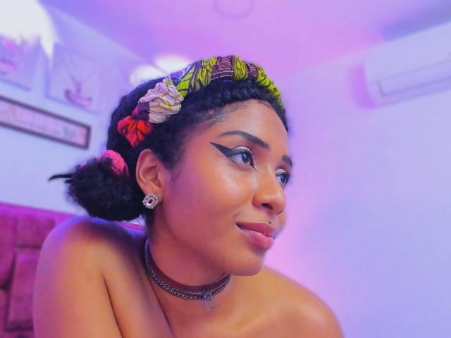 MeganWilliams with dark curly hair, green leaf headband, sharp black eyeliner, pink lipstick, nose ring, choker, earrings, and a confident smile, topless, in a purple-lit room.