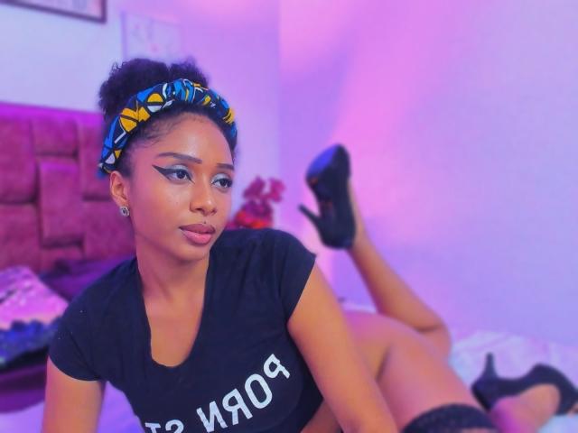 MeganWilliams with dark skin, curly hair in a yellow-blue headband, black t-shirt, and black high heels, lying on a bed, showing her butt and legs raised.