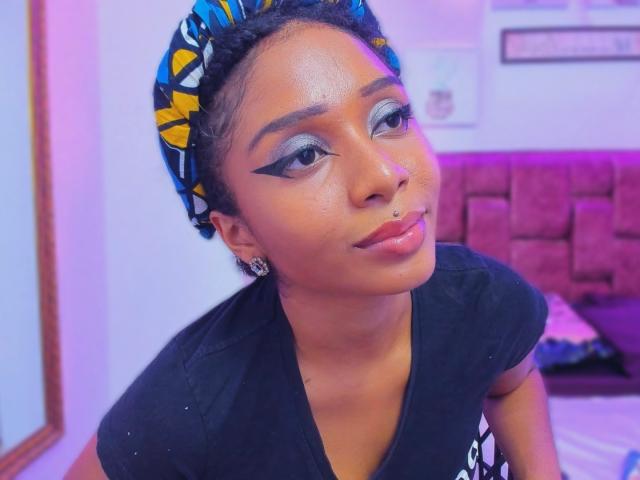 MeganWilliams with dark skin, braided hair in a blue-yellow patterned headscarf, black eyeliner, nose piercing, glossy pink lips, wearing a black shirt, looking up with a slight smile.