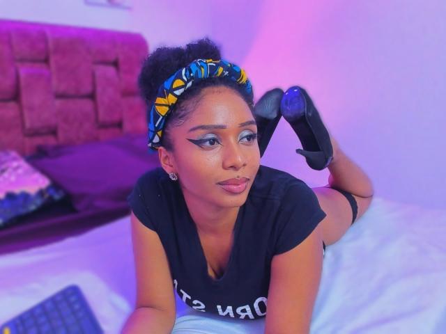 MeganWilliams, laying on bed, black t-shirt, blue and yellow headband, black high heels, butt up, looking at camera, purple and pink lighting, dark skin, makeup, earrings.