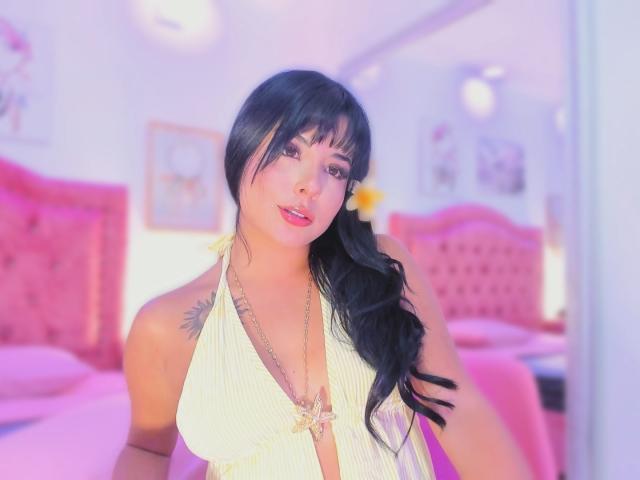GraceClarke with black hair, bangs, and a yellow halter top showing her tits, a tattoo on her shoulder, and a yellow flower in her hair, smiling. Pink bedroom.
